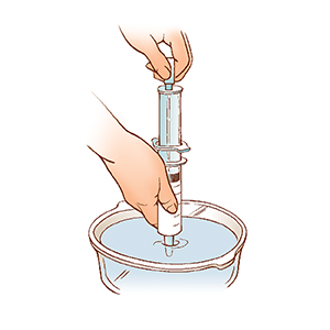 Close-up of hands holding syringe with tip in bowl of water, pulling plunger to draw water into syringe.