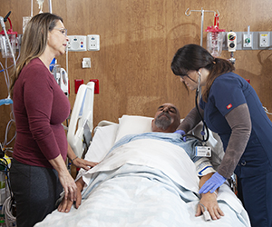 Healthcare provider caring for man in intensive care unit bed while woman stands nearby.